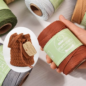 May include: A brown crocheted drawstring bag with a matching tag, displayed with spools of various colored yarn. One spool of brown yarn is labeled "craftl crafty: Soft Crochet". The bag has a textured pattern and a leather drawstring.