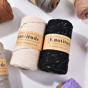 Può includere: Assortimento di rocchetti di cordoncino per macramè in vari colori, tra cui marrone, crema, nero, grigio e rosa. Ogni rocchetto è etichettato con "Knotitude Premium Macrame Cord". Il cordoncino nero ha un effetto glitterato.