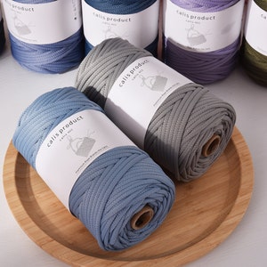 Puede incluir: Bobinas de hilo de macramé azul y gris se muestran en una bandeja de madera. Se ven otras bobinas de hilo de varios colores en el fondo. Cada bobina tiene una etiqueta que dice "calis product".