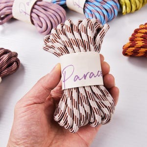 Puede incluir: Una mano sostiene un manojo de paracord beige y marrón, etiquetado con la palabra "Paracord". Otros manojos de paracord de colores están en el fondo. El paracord está bien enrollado y envuelto con una banda de papel.