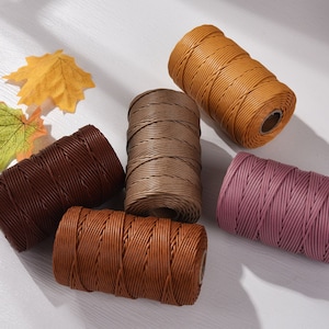 Può includere: Bobine di cordoncino cerato in vari colori, tra cui marrone, beige, arancione e malva. Il cordoncino è avvolto strettamente su anime di cartone. Foglie artificiali gialle e verdi sullo sfondo.