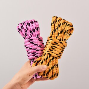 May include: Two bundles of rope, one pink and one yellow, each with a black diamond pattern. The ropes are held together, showcasing their contrasting colors and textures. The image is set against a plain background.