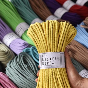 Puede incluir: Un manojo de cuerda de algodón amarilla, con la etiqueta "Hi Basket Rope 6.0", se sostiene frente a una variedad de carretes de cuerda de colores. El fondo presenta carretes de cuerda verde, azul, morado y marrón.