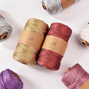 Puede incluir: Surtido de coloridas bobinas de cordón de macramé en plata, oro, rojo, blanco, morado y rosa. Cada bobina está envuelta en una banda de papel marrón con un logotipo. Los cordones están dispuestos sobre una superficie blanca, ideales para manualidades.