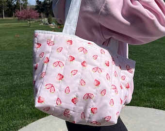 Puede incluir: Una bolsa de mano acolchada de color rosa con una correa blanca y un estampado de fresas. La bolsa está hecha de tela y tiene un diseño acolchado.