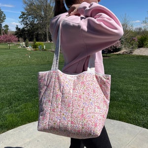 May include: A quilted tote bag with a floral print in shades of pink, white, and green. The bag has white straps and a white lining.