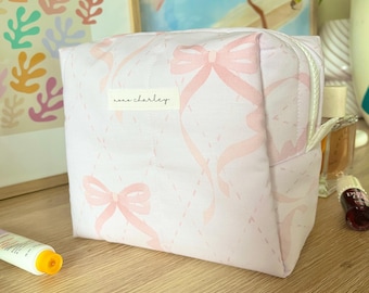 Puede incluir: Un estuche de tela acolchada en color rosa claro con un patrón repetitivo de lazos rosa suave y líneas de diamante discontinuas. Una pequeña etiqueta rectangular blanca en la parte frontal muestra el texto negro "xoxo charley" con una cremallera blanca.