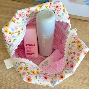 Puede incluir: Un neceser con estampado floral y cremallera blanca, que muestra un interior de cuadros rosas y blancos. Contiene tres productos de belleza: una crema hidratante BYOMA rosa, un producto rhode blanco y un bálsamo labial Glossier rosa.