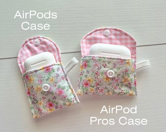 Può includere: Due custodie in tessuto per AirPods. La custodia a sinistra è per AirPods standard e ha un motivo a quadri rosa e bianco all'interno e un motivo floreale all'esterno. La custodia a destra è per AirPods Pro e ha un motivo a quadri rosa e bianco all'interno e un motivo floreale all'esterno. Entrambe le custodie hanno una chiusura a bottone bianco.