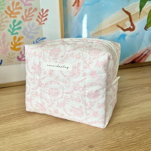 May include: A rectangular fabric pouch featuring a white base with a delicate pink floral pattern. The bag has a zipper closure and a small white rectangular label on the front with the black text "rose charley" printed in a cursive script font.