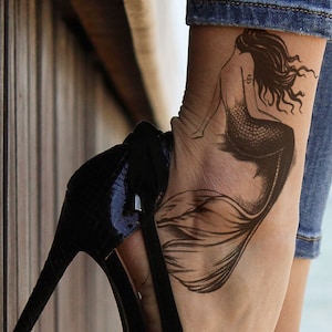 May include: A black high-heeled shoe with a pointed toe and a mermaid tattoo on the ankle. The tattoo is a black and white illustration of a mermaid with long flowing hair.