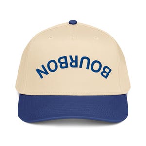Può includere: Un cappellino da baseball con corona color crema e visiera blu navy. La parola "BOURBON" è ricamata in blu navy sulla parte anteriore del cappellino. Il cappellino ha una visiera curva e un piccolo bottone blu navy sulla parte superiore.