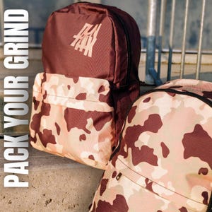 May include: Two camouflage backpacks with a brown and pink color scheme. The larger backpack has a brown top section with the text "PACK YOUR GRIND" in white letters. The smaller backpack has a brown and pink camouflage pattern.