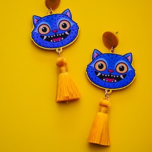 May include: A pair of blue cat-shaped earrings with gold trim, featuring large eyes and a toothy grin. Each earring has a yellow tassel and a brown stud. The background is a solid yellow.
