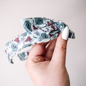 May include: A green headband with a Christmas-themed print featuring a green creature wearing a Santa hat. The headband is tied in a bow.