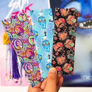 May include: Three decorative bookmarks with colourful patterns are held in front of a poster. The bookmarks feature various designs, including floral patterns with portraits, a blue cat-like creature, and a black background with hearts and illustrations.
