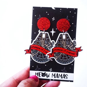 May include: A pair of red glitter earrings featuring a black and white illustration of the Millennium Falcon spaceship from Star Wars. The earrings have a red ribbon with the text "Punch it Chewie" on them. The earrings are displayed on a black card with the text "Meow Mamas".