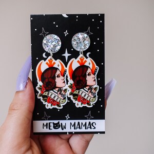 May include: A pair of dangle earrings featuring a cartoon image of Princess Leia with the word "Rebel" printed on the earrings. The earrings are on a black card with white stars and the words "Meow Mamas".