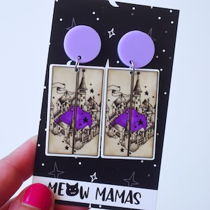 May include: A pair of purple and white earrings featuring a map of a castle with stars. The earrings are on a black card with white polka dots and the text "MEOW MAMAS".