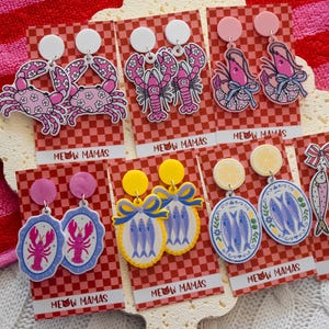 May include: A selection of vibrant acrylic earrings showcasing marine life and food. Designs include crabs, lobsters, shrimp, fish, and sardines. Each pair is presented on a red and white chequered card with the text "MEOW MAMAS".