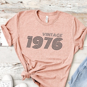 May include: A peach-colored t-shirt with the text "Vintage 1976" in black and white striped font. The shirt is laid flat on a white wood background with a pair of white sneakers and a pair of blue jeans.