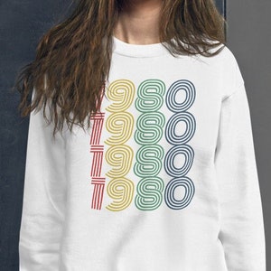 Puede incluir: Sudadera blanca con un gr&aacute;fico colorido del a&ntilde;o 1980 repetido cuatro veces.