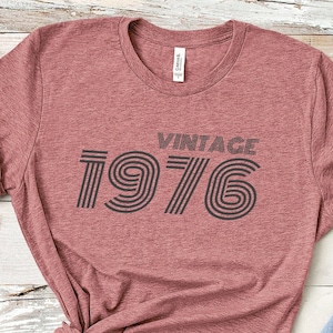 May include: A heather pink t-shirt with a black vintage 1976 graphic printed on the front.