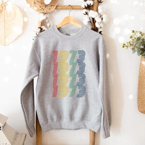 May include: Gray sweatshirt with a rainbow colored graphic of the year 1973 repeated five times.