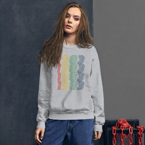 May include: A gray sweatshirt with a retro-style graphic of the year 1983 repeated in a vertical stack. The numbers are outlined in different colors, including yellow, green, blue, and red.
