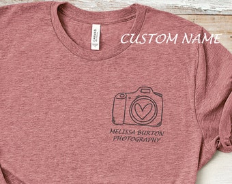 May include: A heather pink t-shirt with a black and white illustration of a camera with a heart in the lens. The text "Melissa Burton Photography" is printed below the camera.