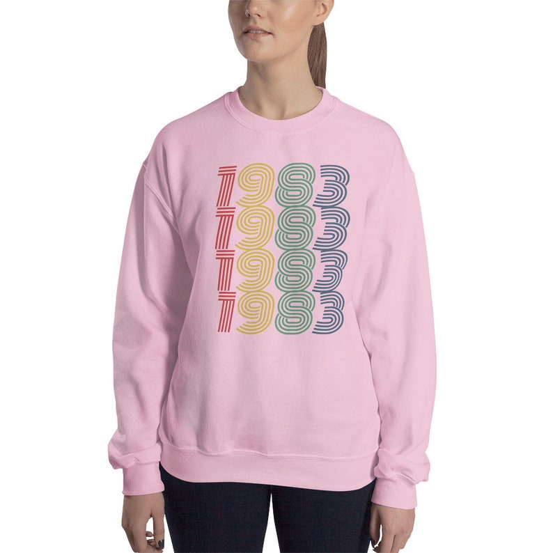 May include: A pink sweatshirt with a retro-style graphic of the year 1983 repeated four times in different colors.