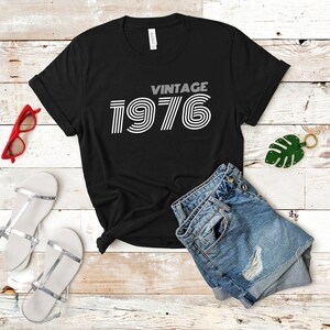 May include: Black t-shirt with white text that reads "Vintage 1976" in a retro font style. The shirt is laid flat on a white wooden surface with a pair of denim shorts, a pair of silver sandals, and a green leaf keychain.