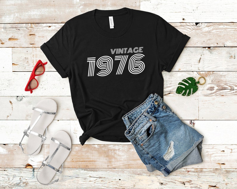 May include: Black t-shirt with white text that reads "Vintage 1976" in a retro font style. The shirt is laid flat on a white wooden surface with a pair of denim shorts, a pair of silver sandals, and a green leaf keychain.