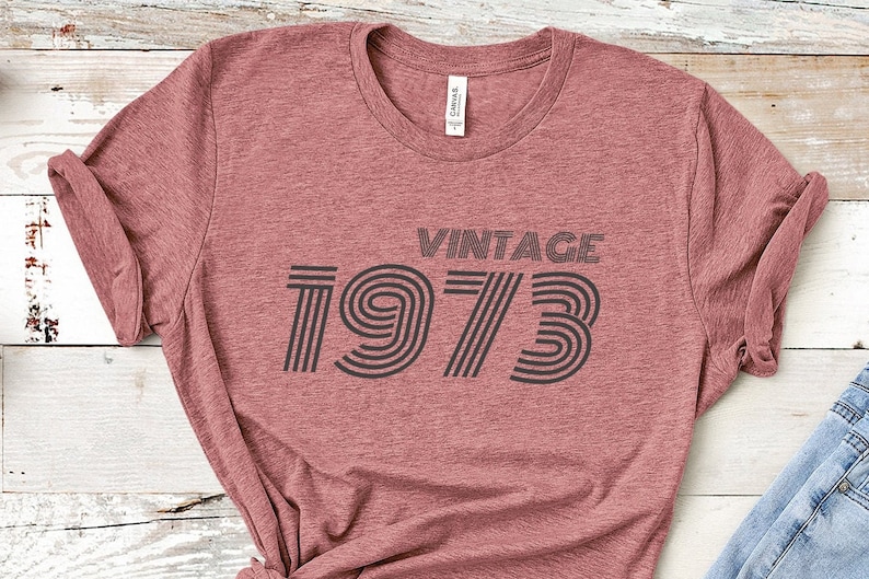 K&ouml;nnte beinhalten: Ein maronfarbenes T-Shirt mit dem Aufdruck "Vintage 1973" in schwarzer Schrift mit einer Retro-Schriftart.