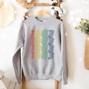 May include: A gray sweatshirt with a colorful graphic design of the year 1983. The numbers are stacked vertically and are outlined in red, yellow, green, and blue.
