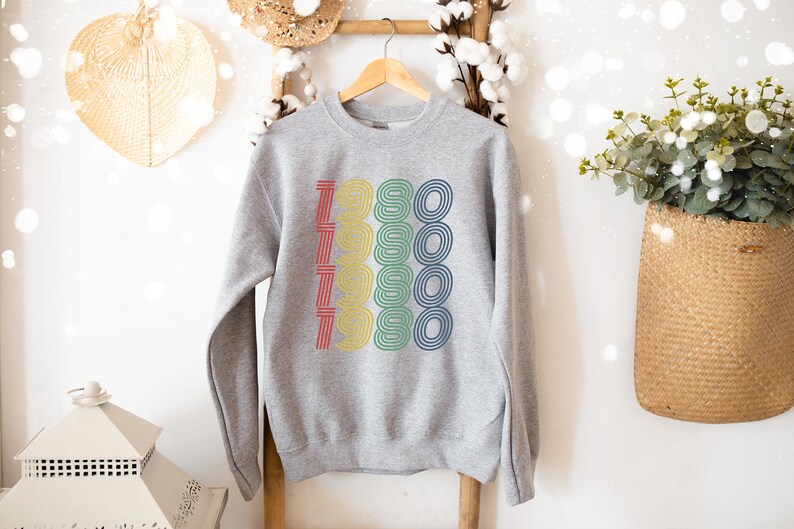 Puede incluir: Una sudadera gris con un gr&aacute;fico retro del n&uacute;mero 1980 repetido cinco veces en diferentes colores. Los colores son rojo, amarillo, verde y azul.