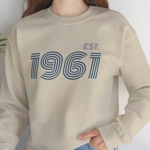 May include: A beige sweatshirt with a blue and white graphic print of the year 1961 with the text "EST." above it.