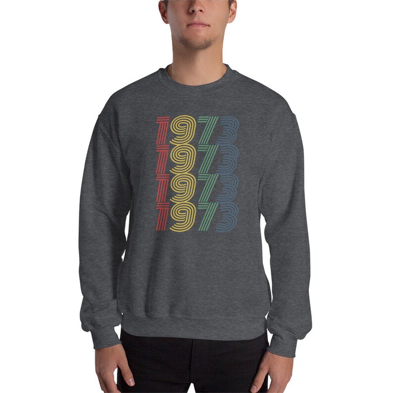 May include: A dark grey sweatshirt with a retro-style graphic of the year 1973 repeated four times in different colors: red, yellow, green, and blue.