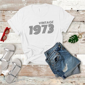 K&ouml;nnte beinhalten: Wei&szlig;es T-Shirt mit dem Text "Vintage 1973" in einer Retro-Schrift. Das Shirt liegt auf einer Holzoberfl&auml;che mit einem Paar silbernen Sandalen, einer roten Sonnenbrille und einer blauen Jeans-Shorts.