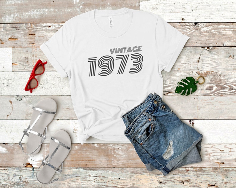 K&ouml;nnte beinhalten: Wei&szlig;es T-Shirt mit dem Text "Vintage 1973" in einer Retro-Schrift. Das Shirt liegt auf einer Holzoberfl&auml;che mit einem Paar silbernen Sandalen, einer roten Sonnenbrille und einer blauen Jeans-Shorts.