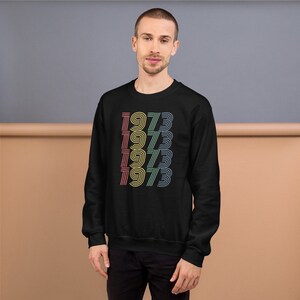 May include: Black sweatshirt with a retro design featuring the year 1973 repeated in a stacked pattern with rainbow colors.