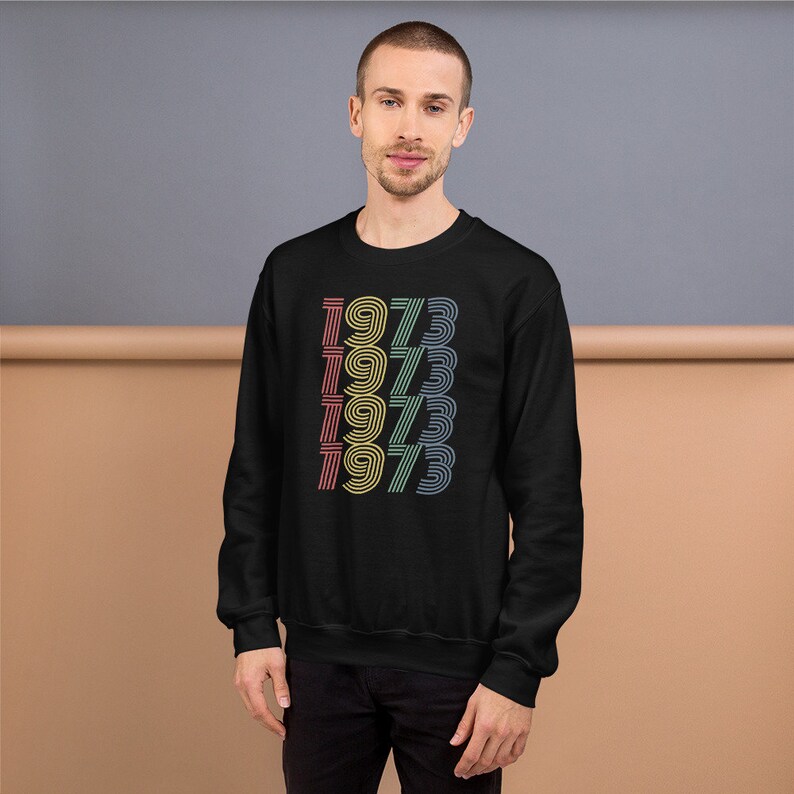 May include: Black sweatshirt with a retro design featuring the year 1973 repeated in a stacked pattern with rainbow colors.