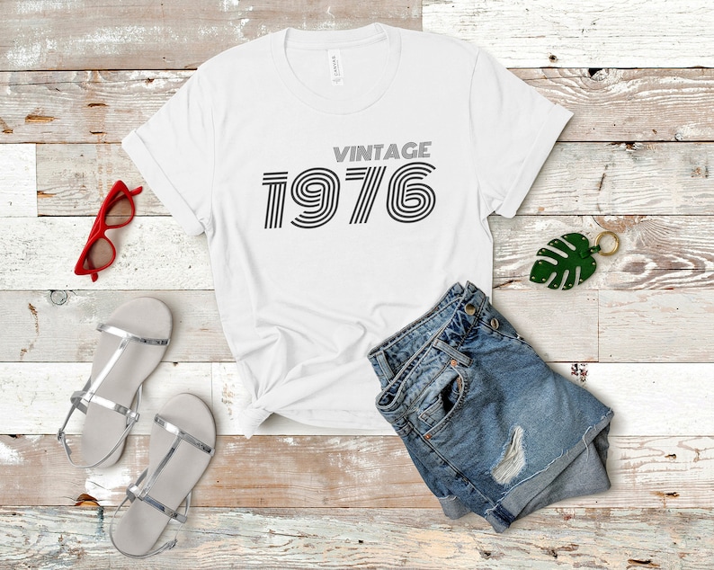 May include: White t-shirt with the text "Vintage 1976" in a retro font. The shirt is laid flat on a wooden surface with a pair of silver sandals, a pair of denim shorts, and a green leaf keychain.
