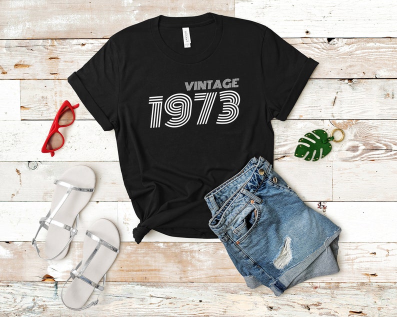 K&ouml;nnte beinhalten: Schwarzes T-Shirt mit wei&szlig;em Text, der "Vintage 1973" lautet. Der Text ist in einem Retro-Schriftart-Stil.