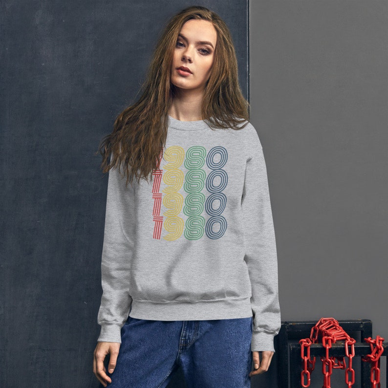 Puede incluir: Una sudadera gris con un gr&aacute;fico retro del a&ntilde;o 1980 repetido cinco veces en diferentes colores. El gr&aacute;fico est&aacute; centrado en la parte delantera de la sudadera.