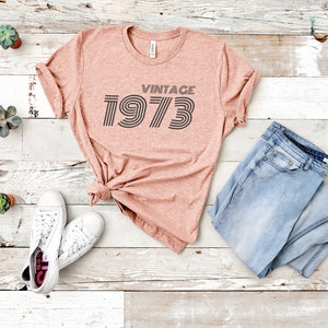 K&ouml;nnte beinhalten: Ein pinkfarbenes T-Shirt mit dem Text "Vintage 1973" in einer Retro-Schriftart. Das Shirt liegt auf einer wei&szlig;en Holzoberfl&auml;che mit einem Paar wei&szlig;en Sneakers und einer blauen Jeans.
