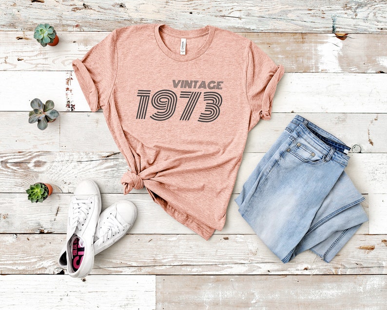 K&ouml;nnte beinhalten: Ein pinkfarbenes T-Shirt mit dem Text "Vintage 1973" in einer Retro-Schriftart. Das Shirt liegt auf einer wei&szlig;en Holzoberfl&auml;che mit einem Paar wei&szlig;en Sneakers und einer blauen Jeans.