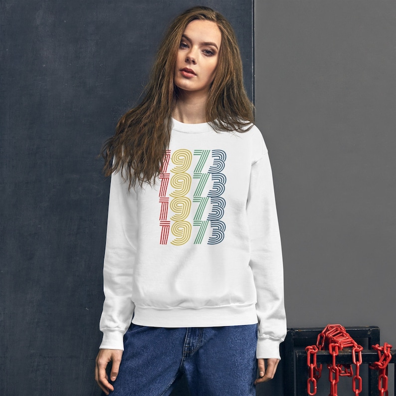 May include: White sweatshirt with a vintage-style graphic of the year 1973 repeated in a rainbow of colors.