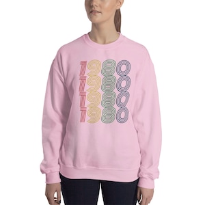 Puede incluir: Sudadera rosa con un dise&ntilde;o retro que presenta el a&ntilde;o 1980 repetido cuatro veces en diferentes colores.