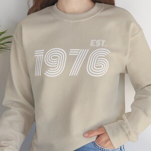 Può includere: Una felpa beige con una stampa grafica bianca dell'anno 1976 con il testo "EST." sopra.